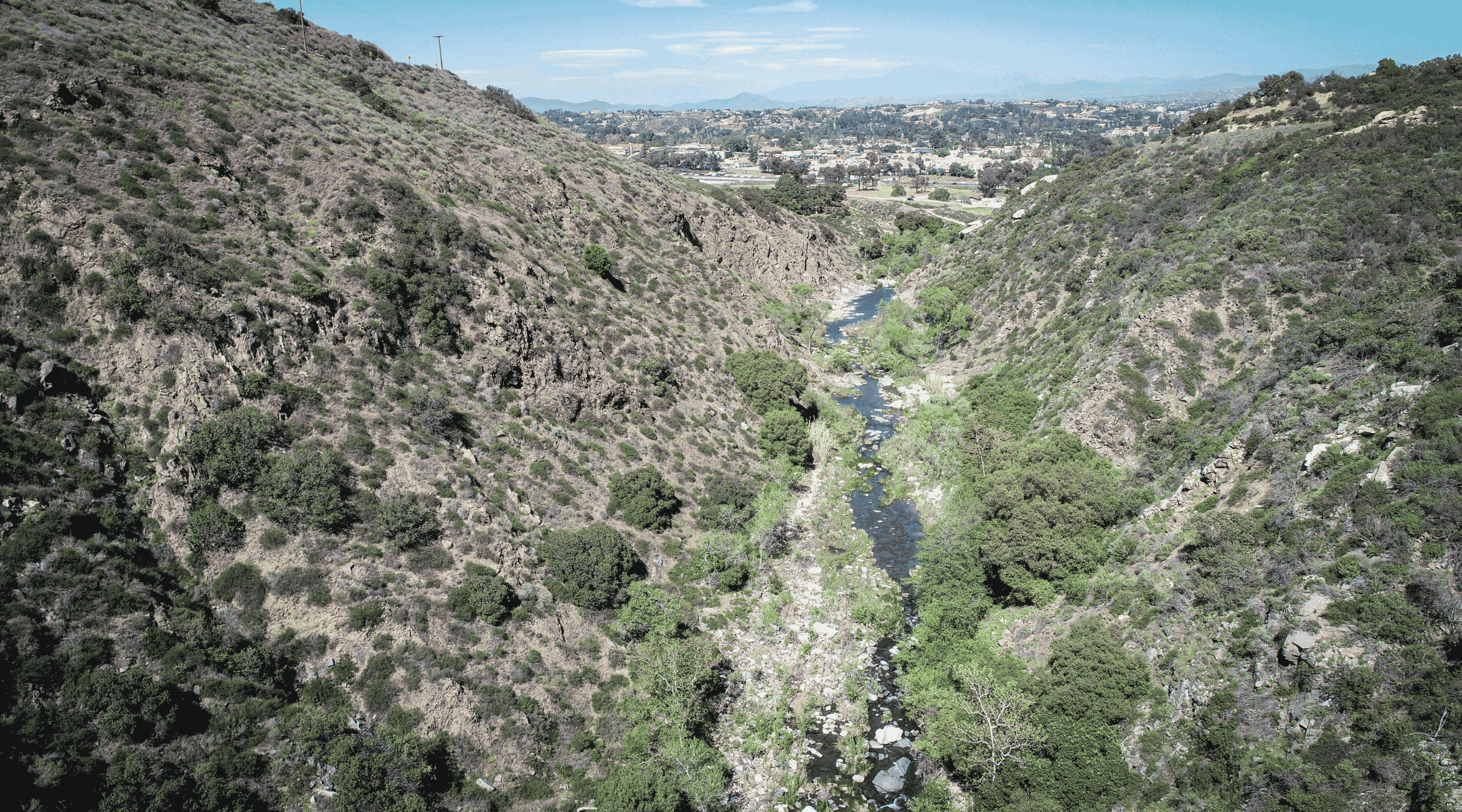 A turquoise stream flows through scrubby hills and and an urban center can be seen in the background