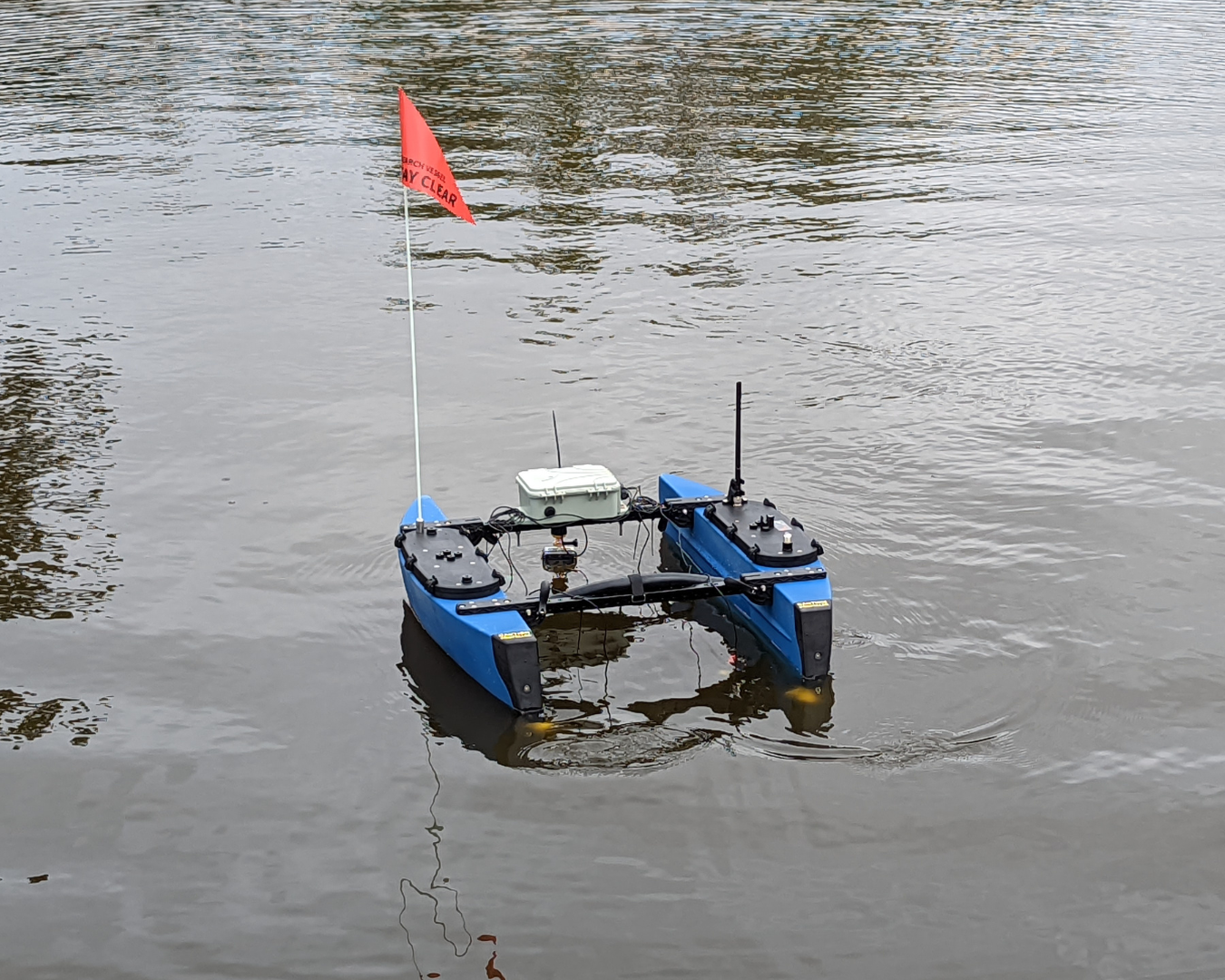 BlueBoat unmanned surface vehicle with twin blue hulls, sensors, antennas, and a red flag floating on lagoon water