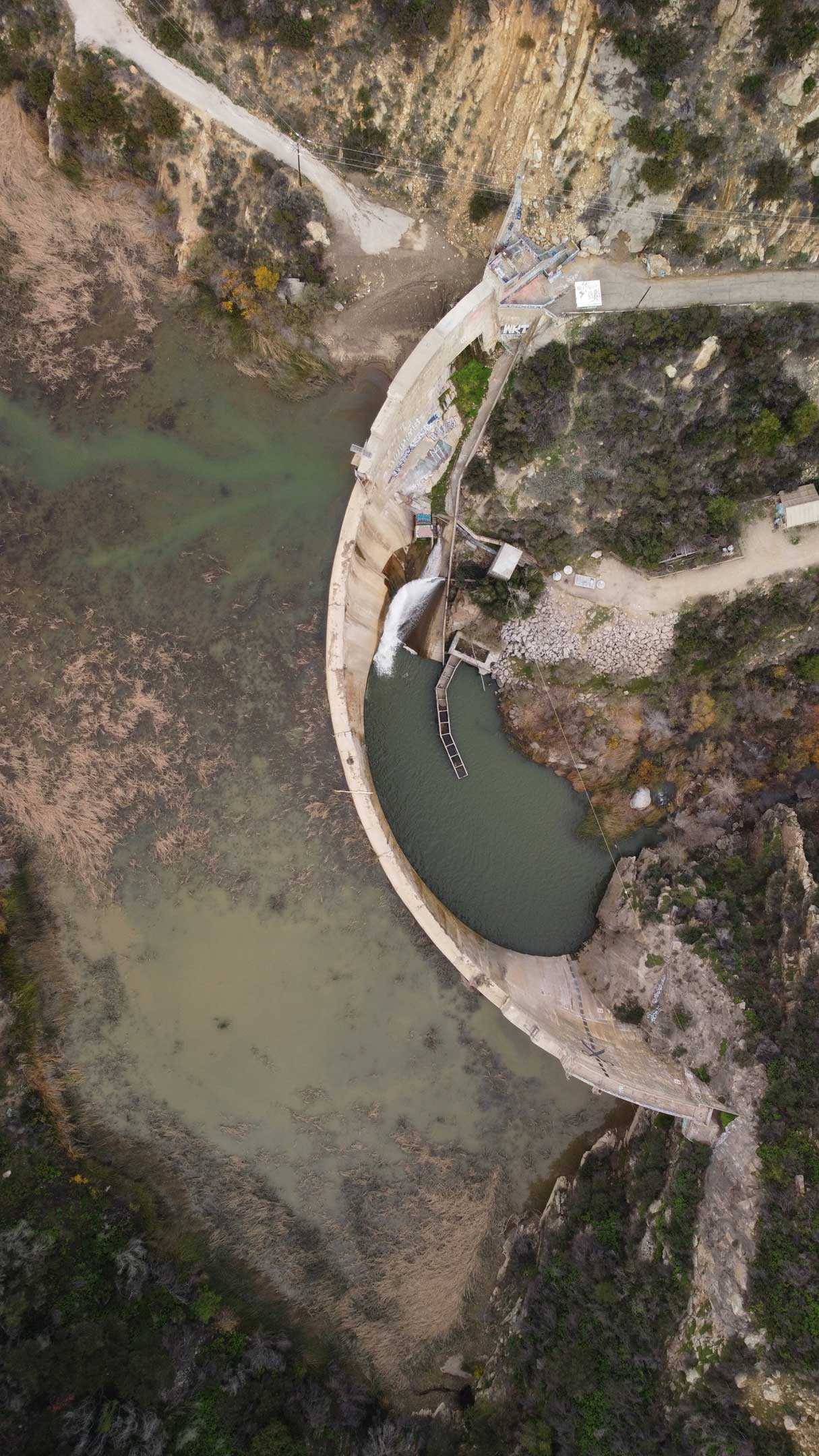 Aerial view of a concrete dam and spillway holding back a river in a rugged, rocky canyon